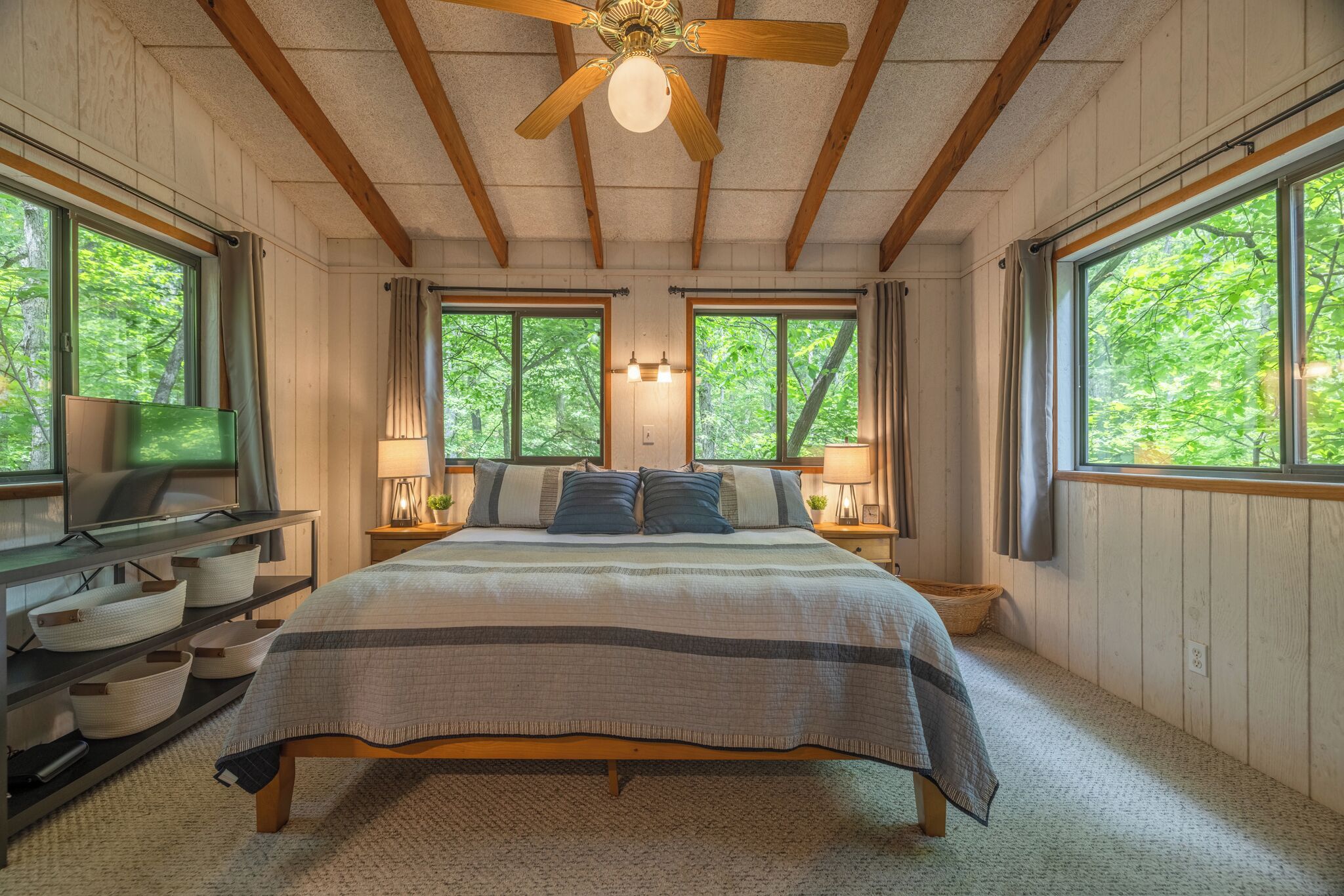 A bedroom with a bed, wooden ceiling beams, a ceiling fan, two windows with curtains, bedside lamps, and a TV on a shelf.
