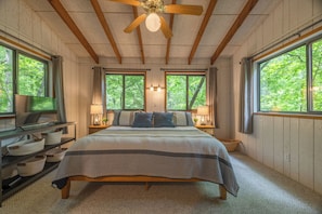 Un dormitorio con cama, vigas de techo de madera, ventilador de techo, dos ventanas con cortinas, lámparas de noche y un televisor sobre una estantería.