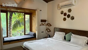 A bedroom with a bed, a window seat, a view of greenery, and a wall-mounted air conditioner. - Stay Leisurely Blue Leaf 3BHK Alibaug (Alibag)
