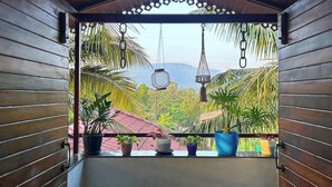 A wooden balcony with a view of a lush landscape, including palm trees and a red-roofed building. - Stay Leisurely Blue Leaf 3BHK Alibaug (Alibag)