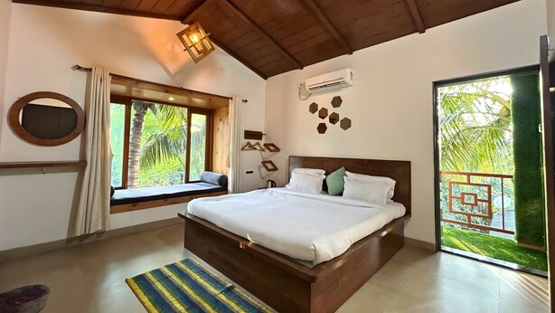A bedroom with a large bed, a window seat, an air conditioning unit, and a view of greenery. - Stay Leisurely Blue Leaf 3BHK Alibaug (Alibag)