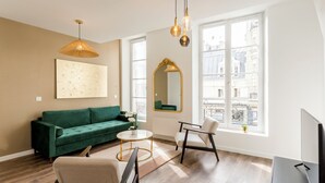 A modern living room with a green sofa, a round coffee table, a white armchair, and a flat-screen TV. There is a large window with a view of buildings, a hanging pendant light, and a framed mirror on the wall. - Merveil Family Suite - Fbg St Antoine (Paris)