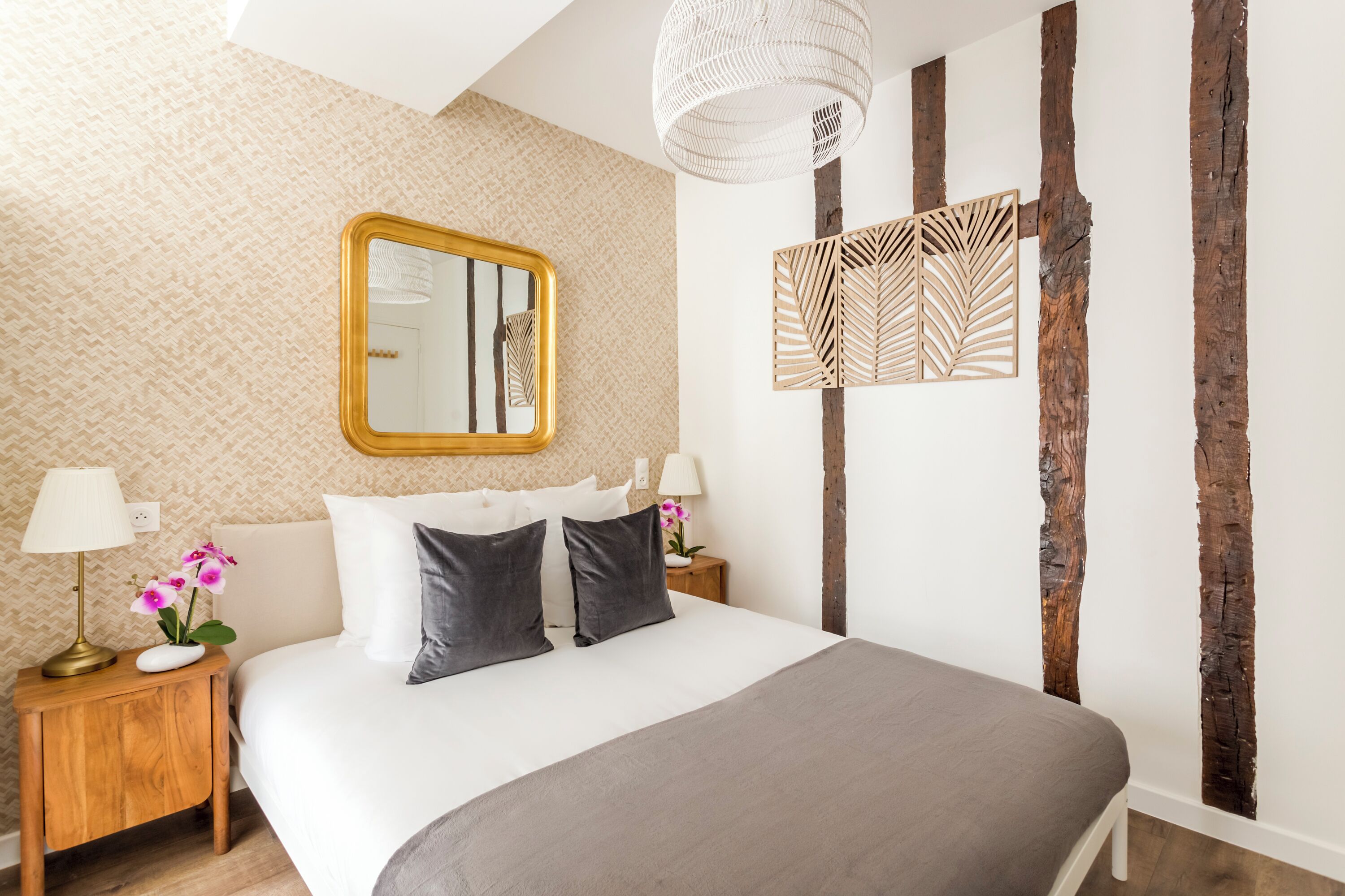A neatly arranged bedroom with a large bed, a wooden nightstand with a lamp, a mirror, and a decorative panel on the wall.