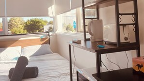 A bedroom with a bed, a desk with a computer, a shelf with kitchen items, and a window with a view of trees. - Shared house in Marsfield-Bright and Sunny/Bus/Mac Uni/Mac Centre (Marsfield)