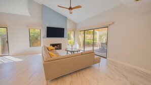 A spacious living room with a beige sofa, a fireplace, a ceiling fan, and large windows offering a view of the outdoors. - Modern Luxury at Ironwood Country Club Palm Desert (Palm Desert)