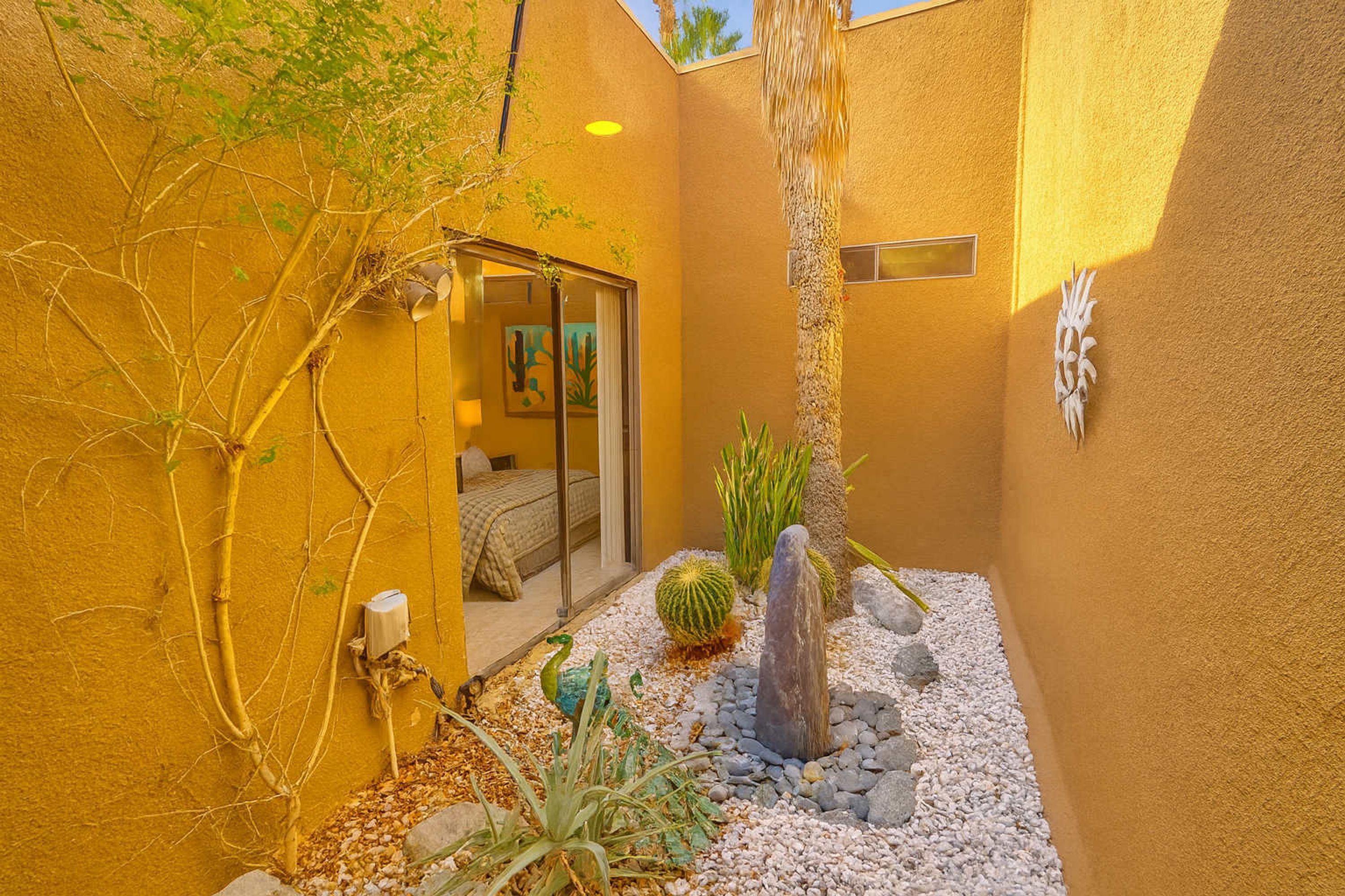 A small, enclosed outdoor area with a yellow wall, a cactus, a rock formation, and a door leading to a room with a bed and a window.