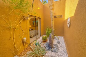 A small, enclosed outdoor area with a yellow wall, a cactus, a rock formation, and a door leading to a room with a bed and a window. - Modern Luxury at Ironwood Country Club Palm Desert (Palm Desert)
