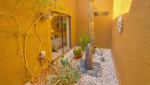 A small, enclosed outdoor area with a yellow wall, a cactus, a rock formation, and a door leading to a room with a bed and a window. - Modern Luxury at Ironwood Country Club Palm Desert (Palm Desert)