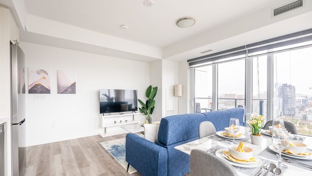A modern living room with a sofa, dining table set for four, a flat-screen TV, and a large window with a city view. - High Rise 1BR Condo w Balcony Beautiful Views (Kitchener)