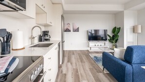 A modern kitchen with a stove, sink, and coffee maker. There is a blue sofa and a TV in the living area. - High Rise 1BR Condo w Balcony Beautiful Views (Kitchener)