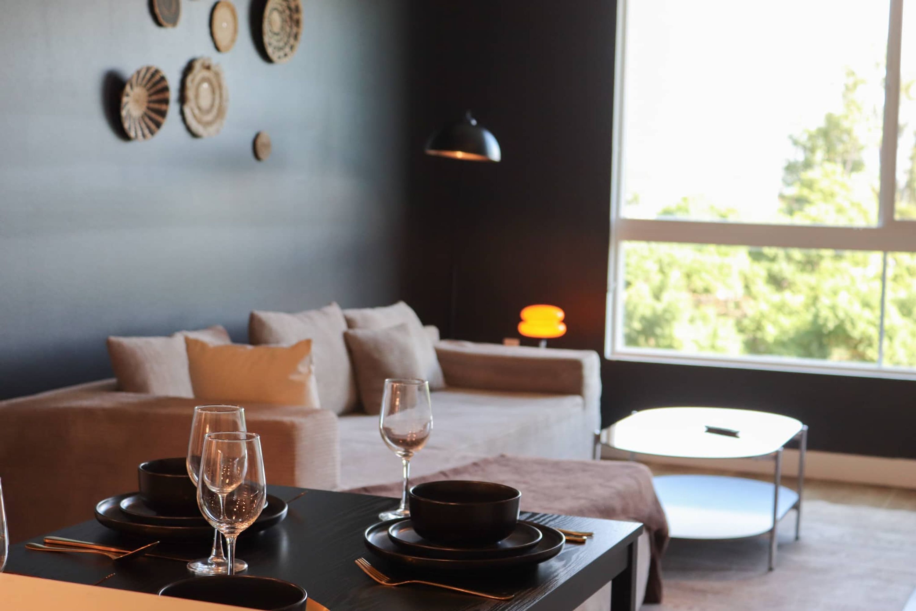 A dining area with a table set for two, a sofa, a lamp, and a view of greenery outside.