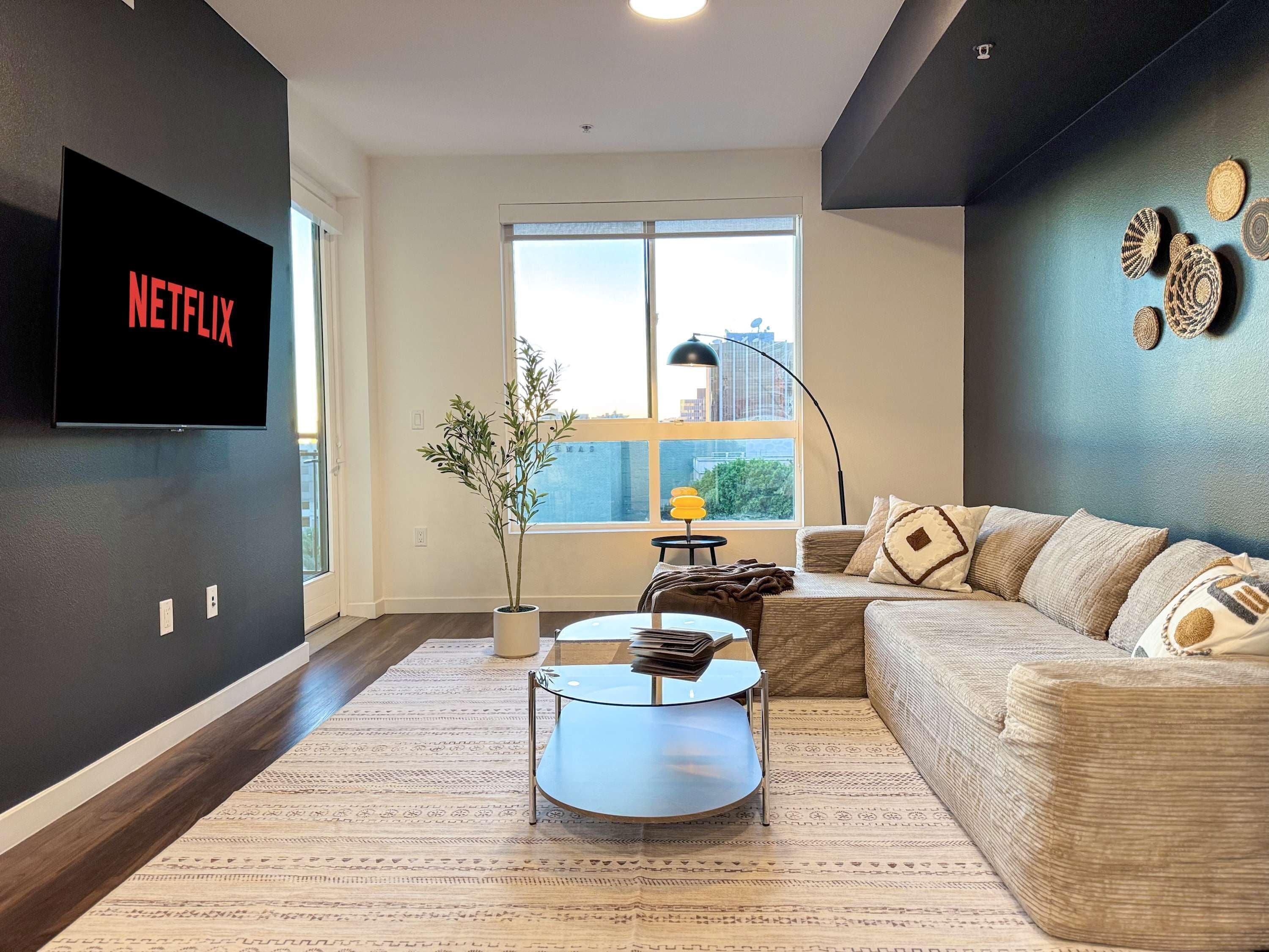 A modern living room with a large TV displaying 'NETFLIX', a sofa, a coffee table, a potted plant, and a view of buildings through a large window.