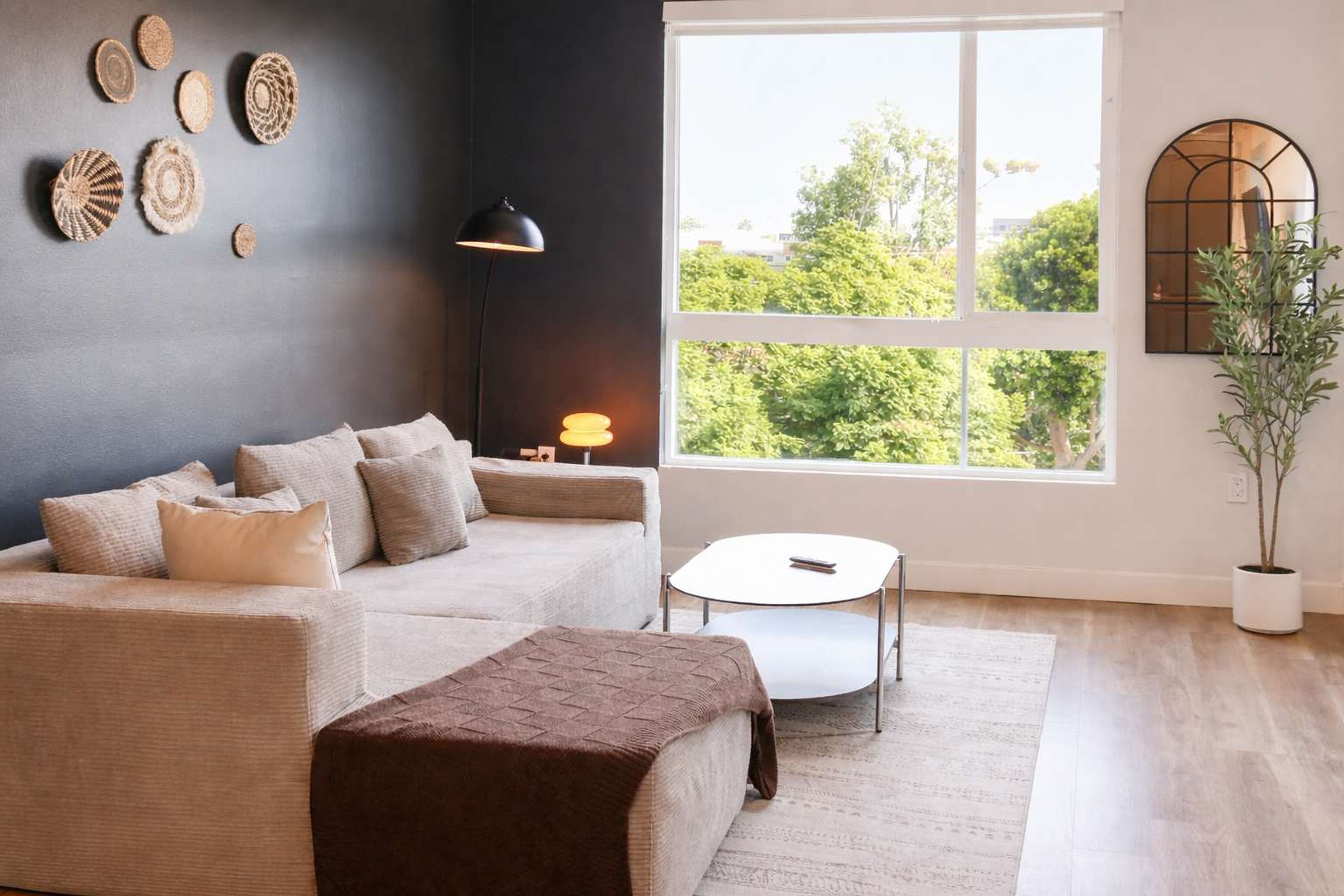 A living room with a sofa, a small table, a lamp, a potted plant, and a large window with a view of trees.