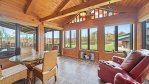 Una habitación amplia con una mesa de vidrio, sillas de mimbre, un sofá de cuero y ventanas grandes que ofrecen vistas al exterior.