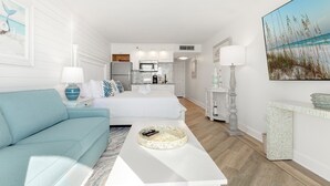 Una habitación moderna y bien iluminada con un sofá azul, una mesa de centro blanca y una cama con cabecera blanca. Se observa una zona de cocina al fondo con armarios blancos y un microondas.