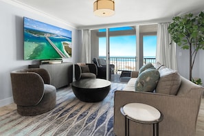 A modern living room with a large flat-screen TV, comfortable seating, a coffee table, and a view of the beach through large windows.