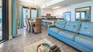 Uma sala de estar espaçosa com um sofá azul, área de jantar com cadeiras de vime e cozinha com armários brancos e eletrodomésticos de aço inoxidável.