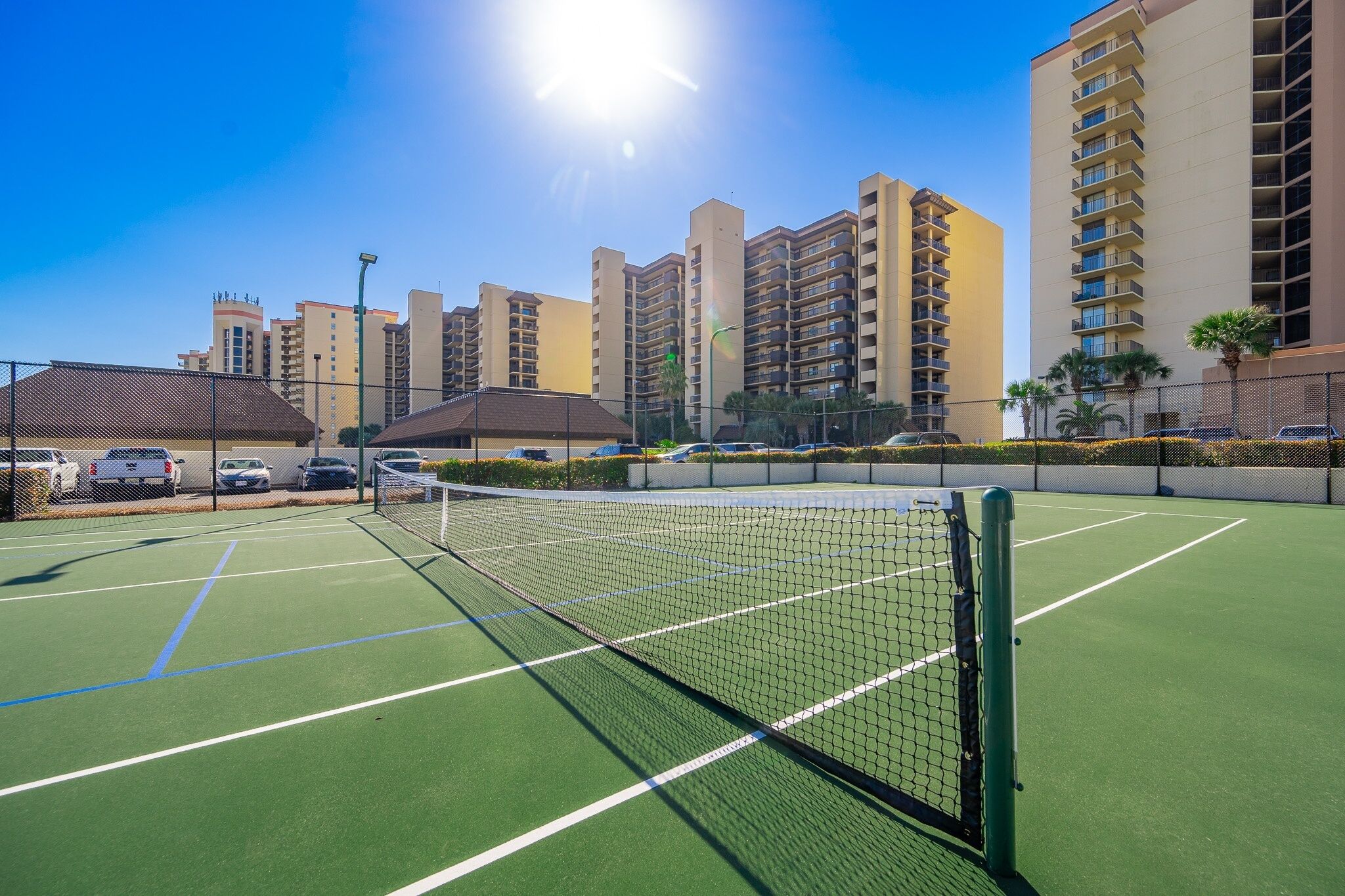 A tennis court with a net, surrounded by a fence, and adjacent to a parking area with cars.
