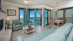 Sala de estar com sofá verde, duas poltronas verdes, mesa de centro de madeira e uma cadeira de vime. Há uma lareira, uma placa com a palavra 'BEACH' e vista para o oceano através de amplas janelas.