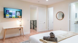 A hotel room with a bed, a desk, a TV displaying streaming services, a mirror, and a door leading to another room.