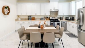 A modern kitchen with white cabinets, a stainless steel refrigerator, a built-in oven, and a microwave. There is a dining table with chairs and a vase with dried plants.