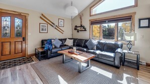 Ein Wohnzimmer mit Holztür, einem schwarzen Ledersofa, einem Couchtisch und einem großen Fenster mit Blick auf Bäume.