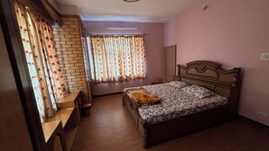 A bedroom with a wooden bed, a small bedside table, a window with floral curtains, and a brick wall.
