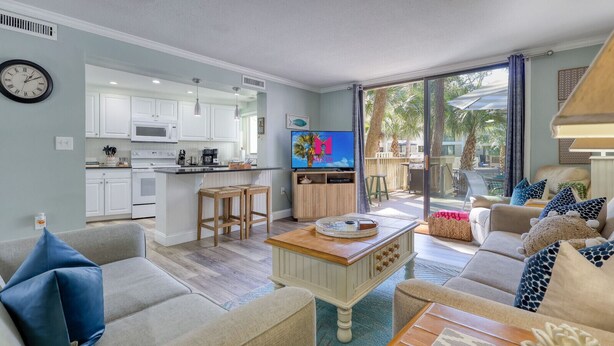 Una sala de estar moderna con un sofá, una mesa de centro y un televisor. Cuenta con una zona de cocina con armarios blancos y un microondas.