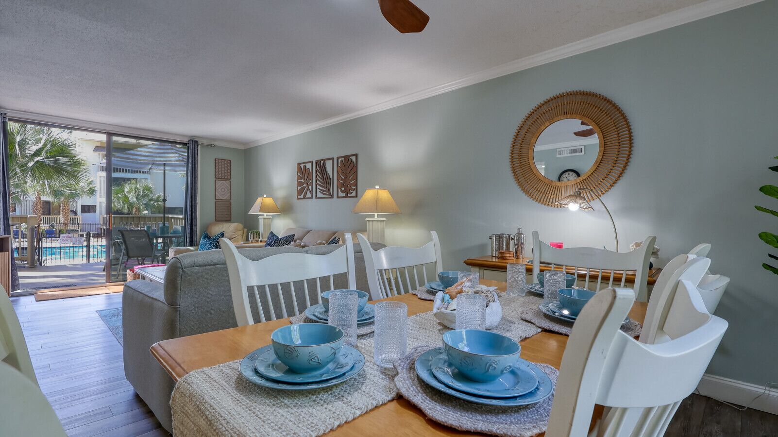 Comedor con una mesa de madera dispuesta para cuatro personas, rodeada de sillas blancas. Hay platos y cuencos azules sobre la mesa, y un espejo redondo cuelga de la pared superior.