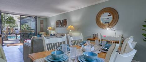 Comedor con una mesa de madera dispuesta para cuatro personas, rodeada de sillas blancas. Hay platos y cuencos azules sobre la mesa, y un espejo redondo cuelga de la pared superior.