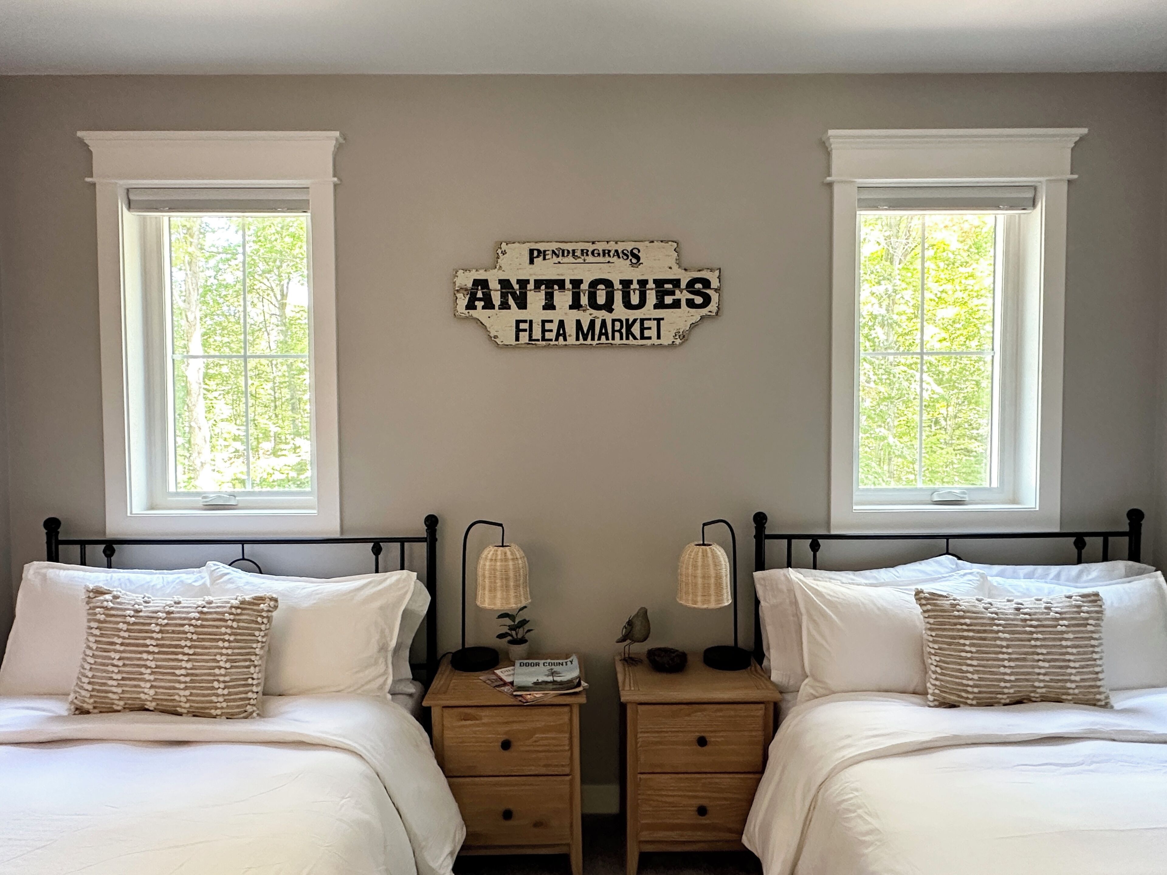 A bedroom with two beds, a wall sign reading "Pendergrass Antiques Flea Market", and windows with a view of greenery.