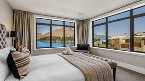 A bedroom with a large bed, two side chairs, and a view of a lake and mountains through large windows.