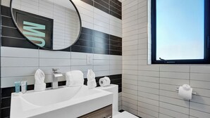 Et moderne badeværelse med en hvid vask, spejl og toilet. Der er håndklæder og toiletartikler på vasken, og et vindue over toilettet.