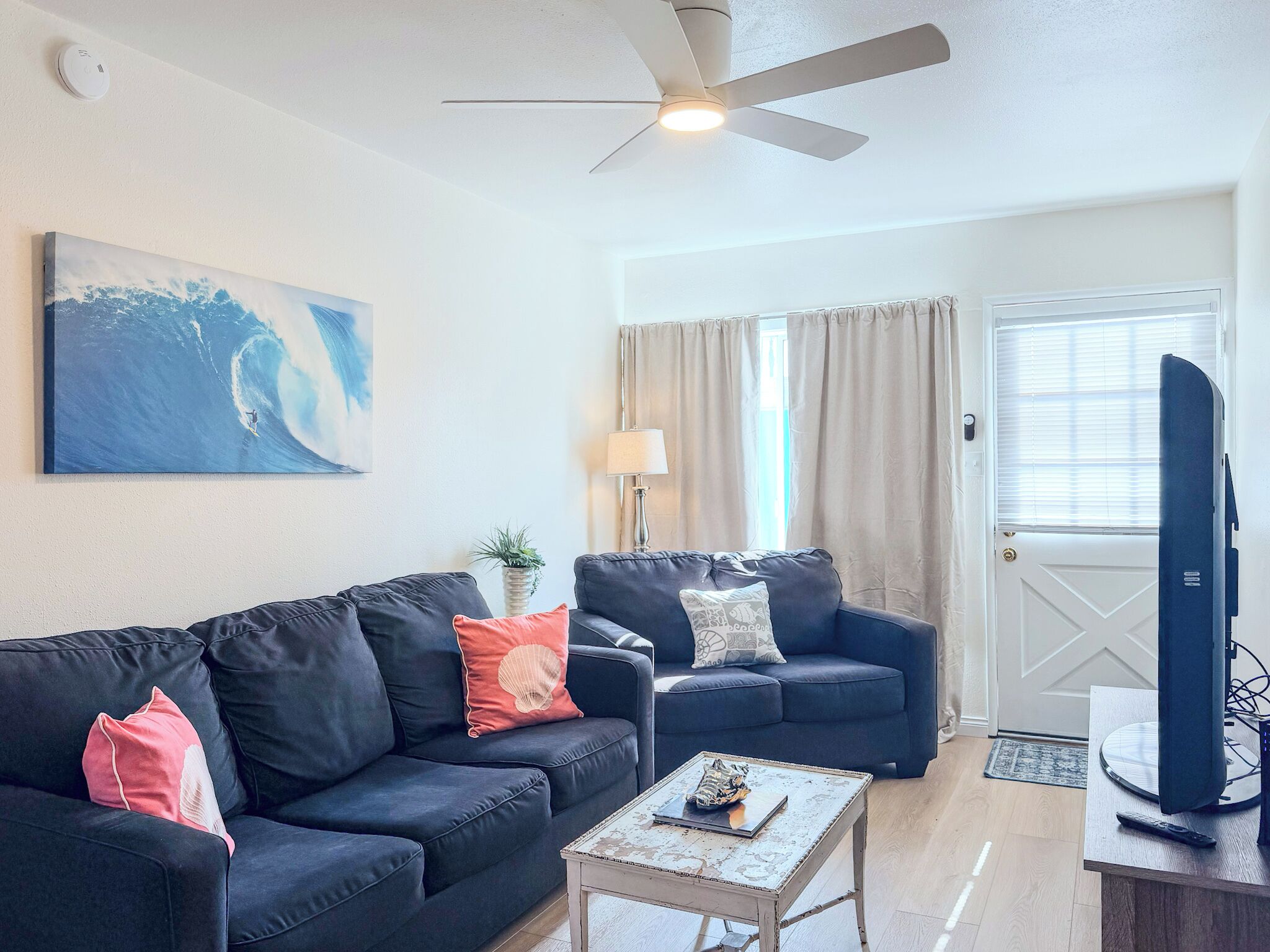 A living room with a dark blue sofa, a smaller sofa, a coffee table, a ceiling fan, a painting of a wave, a lamp, and a door with curtains.