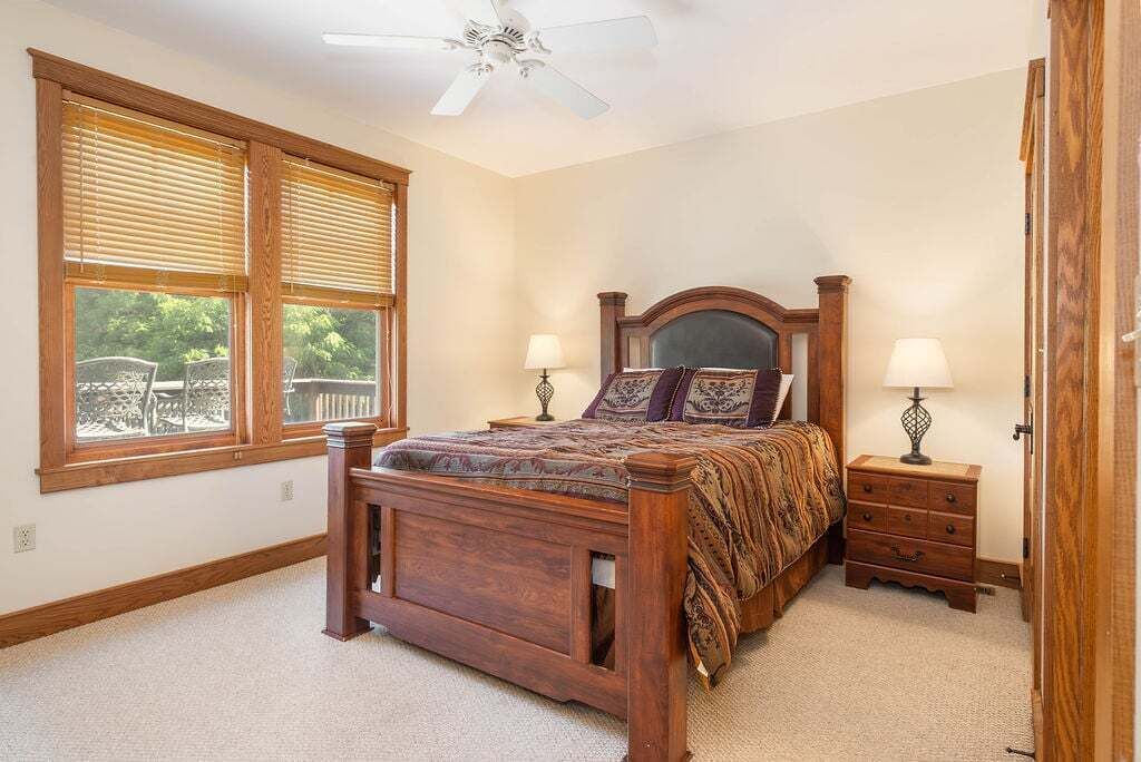 A bedroom with a wooden bed, two bedside tables with lamps, a ceiling fan, and large windows with blinds.