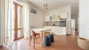 A modern kitchen with white cabinets, a tiled backsplash, and a built-in oven. There is a dining area with a table and chairs, and a large window with curtains. - Gorizia Apartment by Wonderful Italy (Palermo)