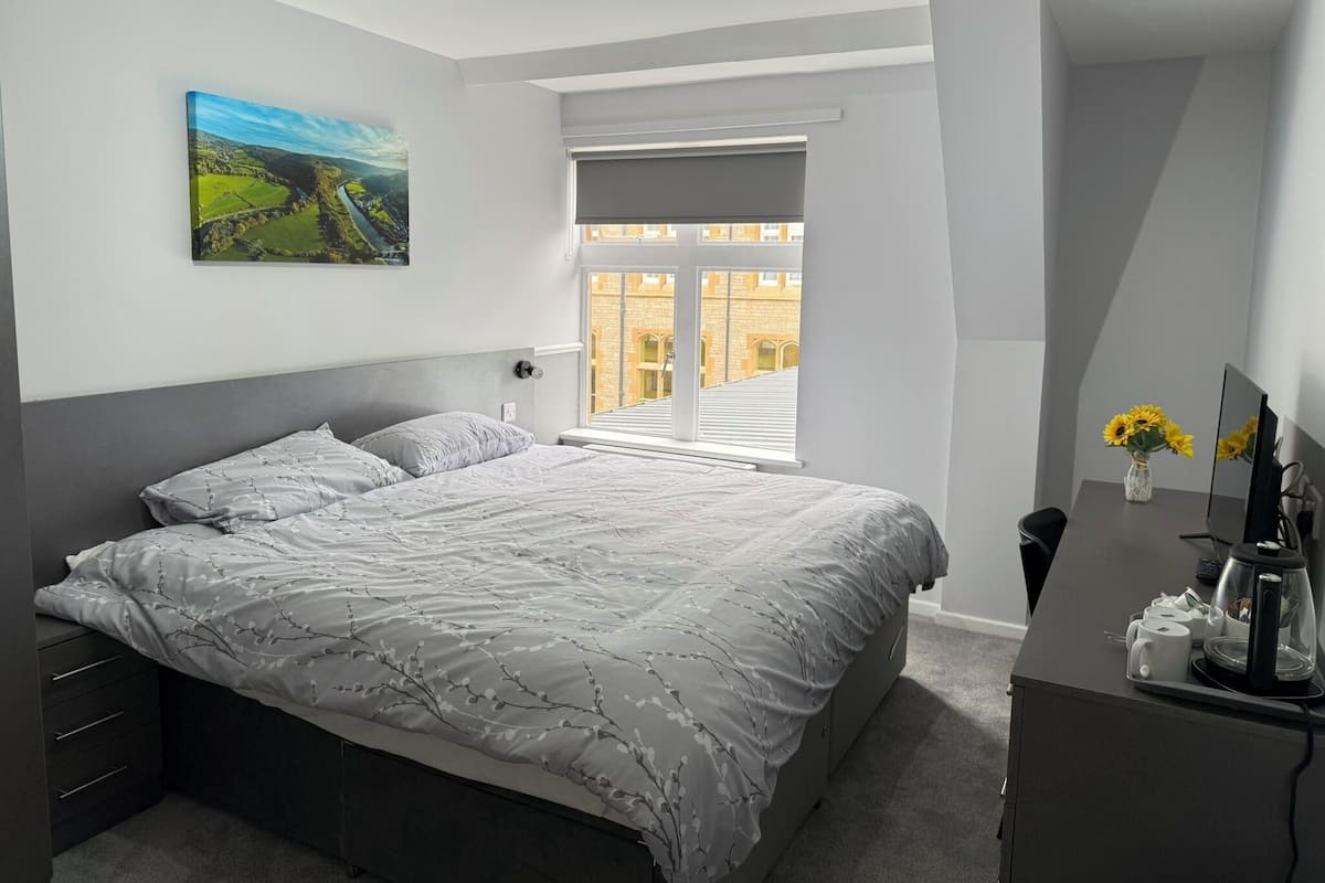 A bedroom with a bed, a desk, a chair, a window, and a painting on the wall.