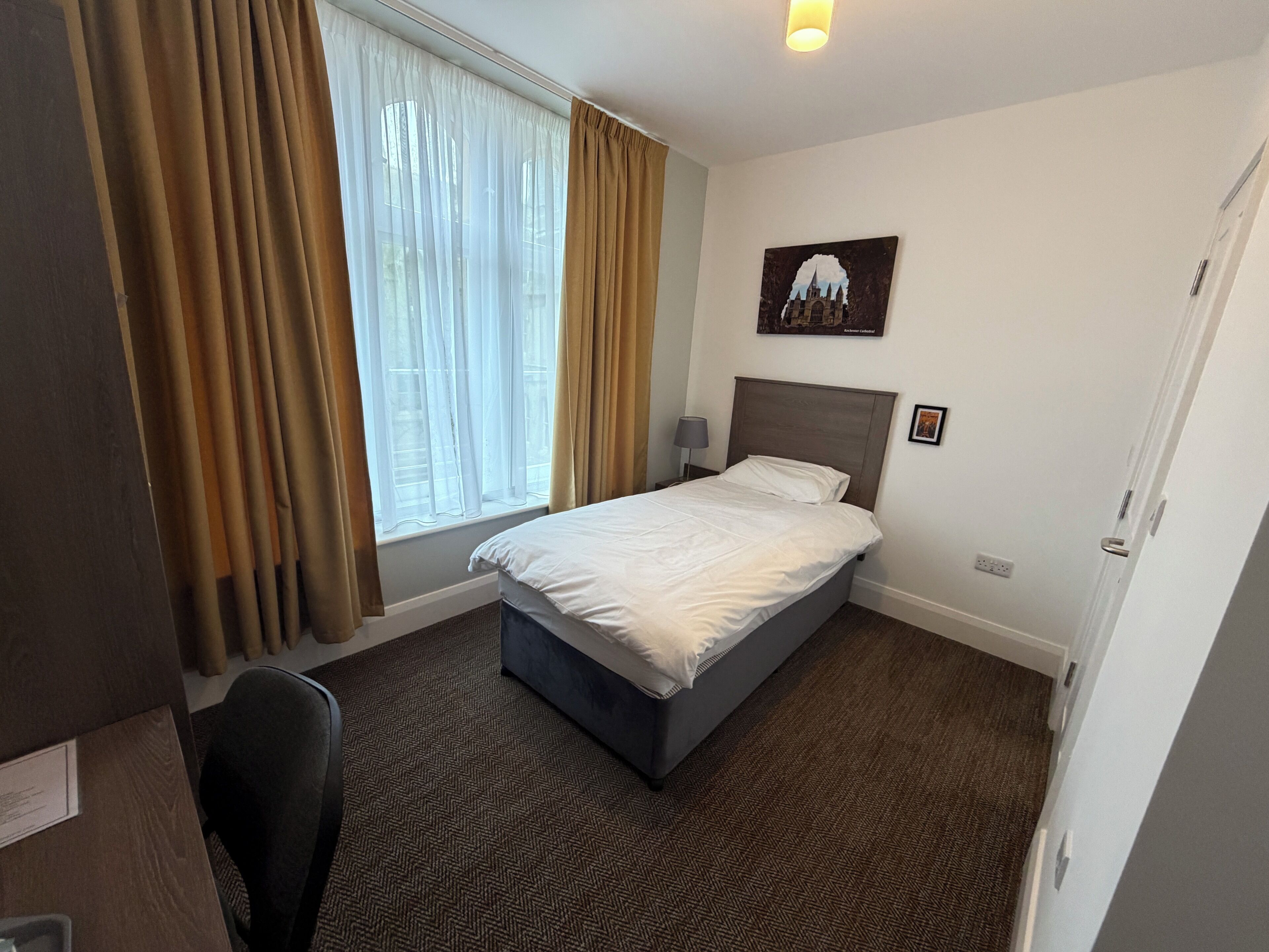 A hotel room with a bed, a desk, a chair, a window with curtains, a nightstand, and a wall-mounted picture.
