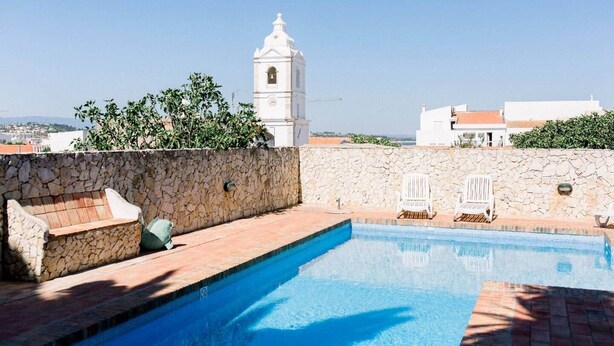 Une piscine sur le toit, avec des murs en pierre, des chaises longues blanches et une tour blanche en arrière-plan.