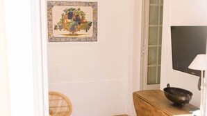 Ein kleiner Raum mit Fliesenboden, einem Holztisch mit einer Schüssel, einem Korbstuhl und einem gerahmten Bild an der Wand.