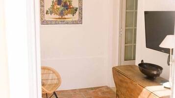 A small room with a tiled floor, a wooden table with a bowl, a wicker chair, and a framed picture on the wall.