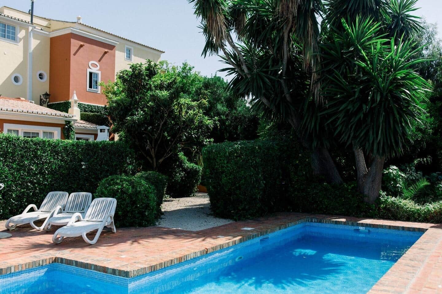 A swimming pool with a tiled edge, surrounded by a brick patio. There are white lounge chairs along the poolside.