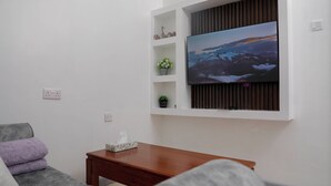 A modern living room with a flat-screen TV mounted on a white wall, a grey sofa, a wooden coffee table, and decorative items on shelves. - Nakuru Guest Suites (Nakuru)