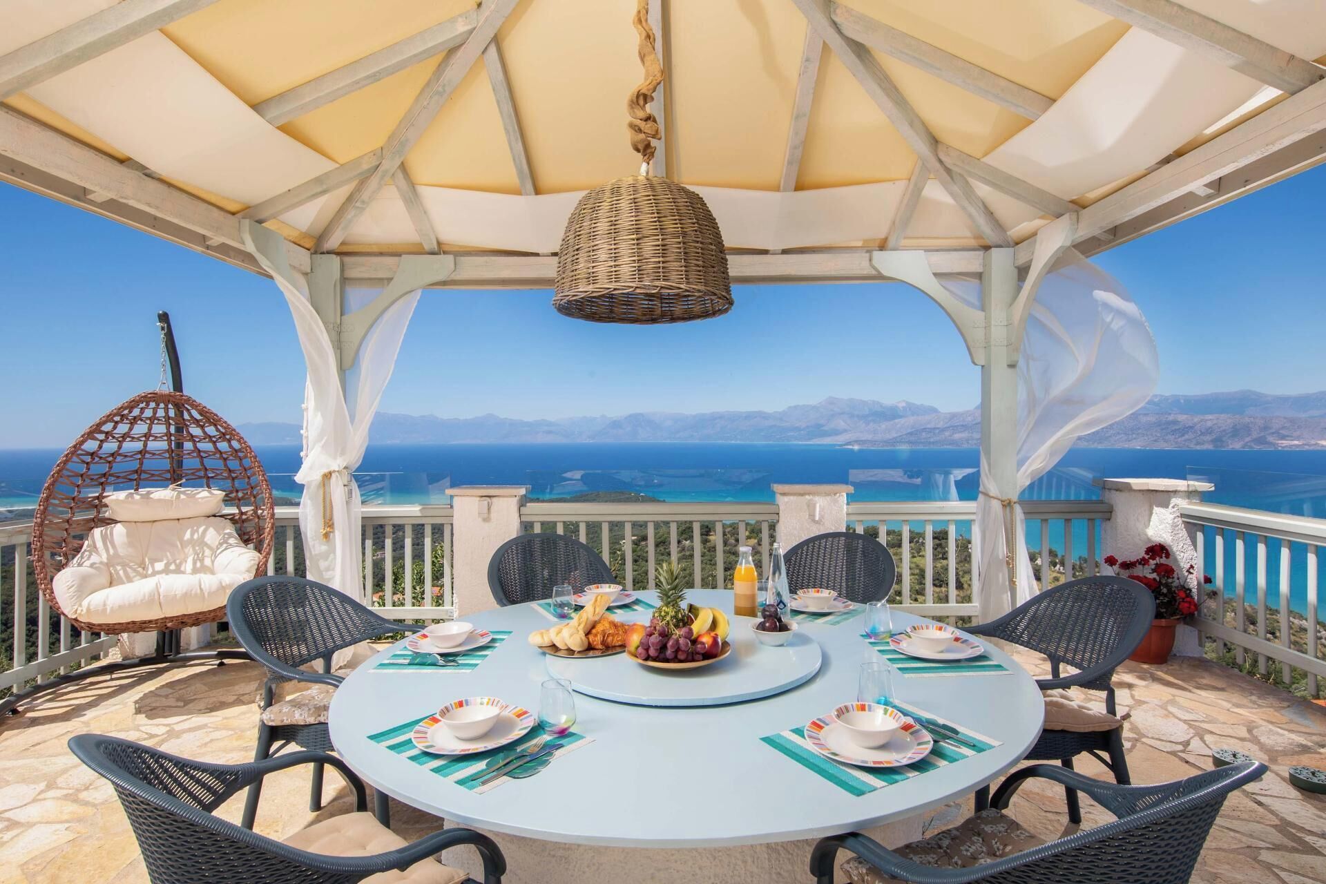 A covered outdoor dining area with a table set for four, surrounded by wicker chairs, overlooking a scenic view of the sea and mountains.