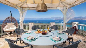 A covered outdoor dining area with a table set for four, surrounded by wicker chairs, overlooking a scenic view of the sea and mountains. - Orizzonte Blu - Three Bedroom Villa, Sleeps 6 (Corfu)