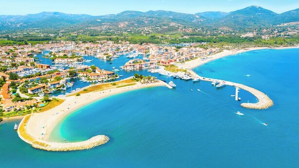 Vista aérea de uma cidade costeira com um porto de iates, praias arenosas e montanhas ao fundo.