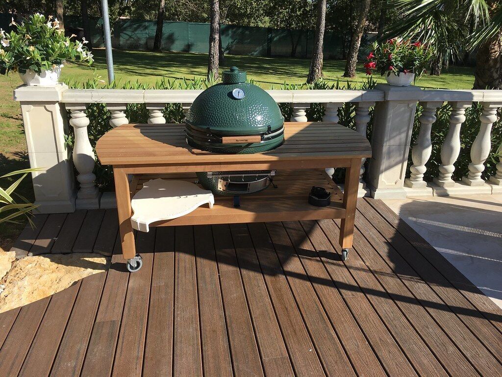A portable grill on a wooden deck with a railing and potted plants in the background.