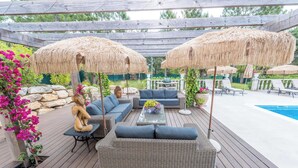 Um deck de madeira com móveis de exterior, incluindo sofás e uma mesa, sob guarda-sóis de palha, e uma piscina ao fundo.