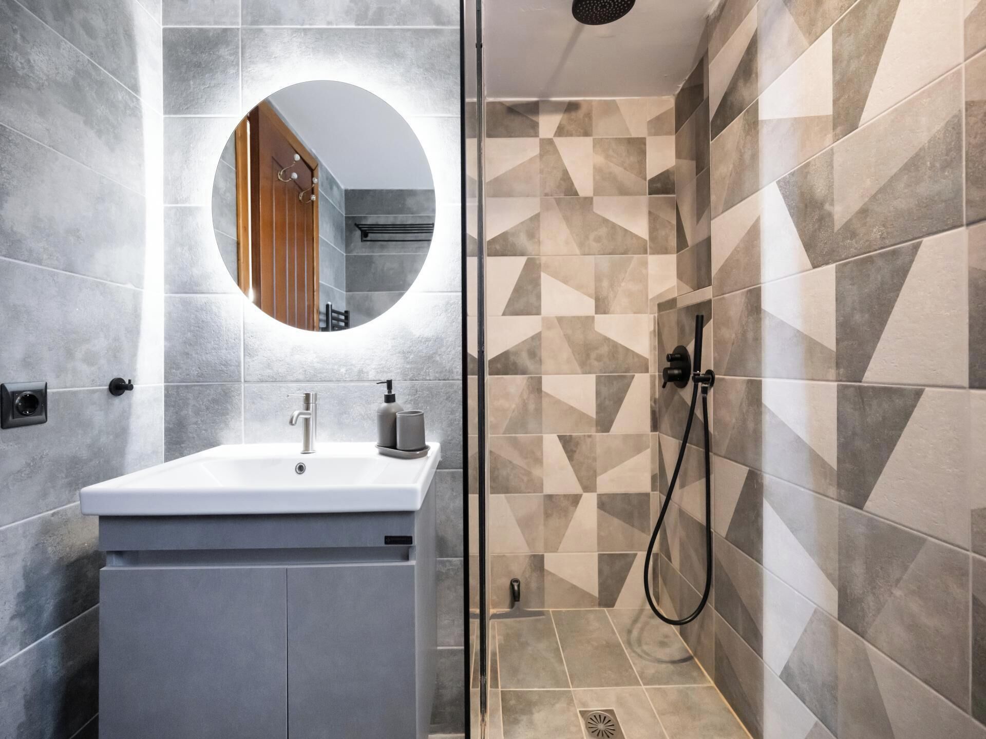 Un baño moderno con un lavamanos blanco, un espejo redondo y una ducha con azulejos de diseño geométrico.