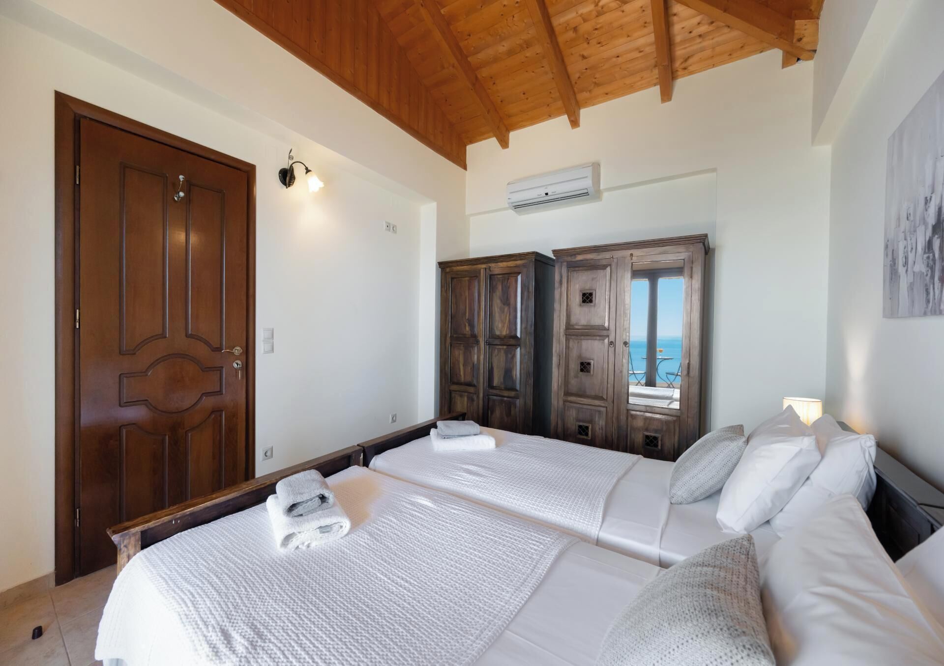 A hotel room with two beds, wooden wardrobes, a view of the sea, and an air conditioning unit.